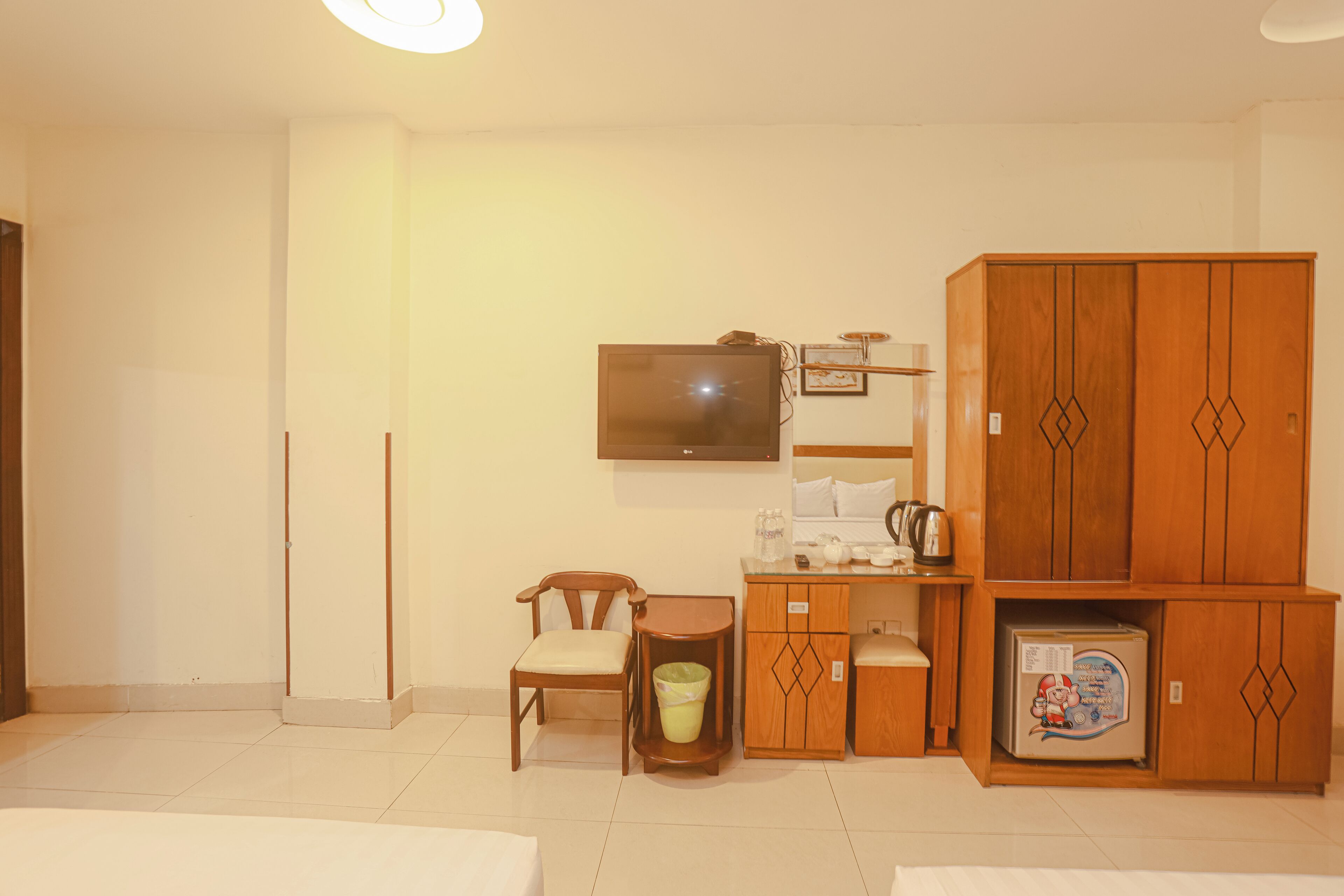A hotel room with a bed, a wooden wardrobe, a television, a small table with a chair, and a refrigerator.