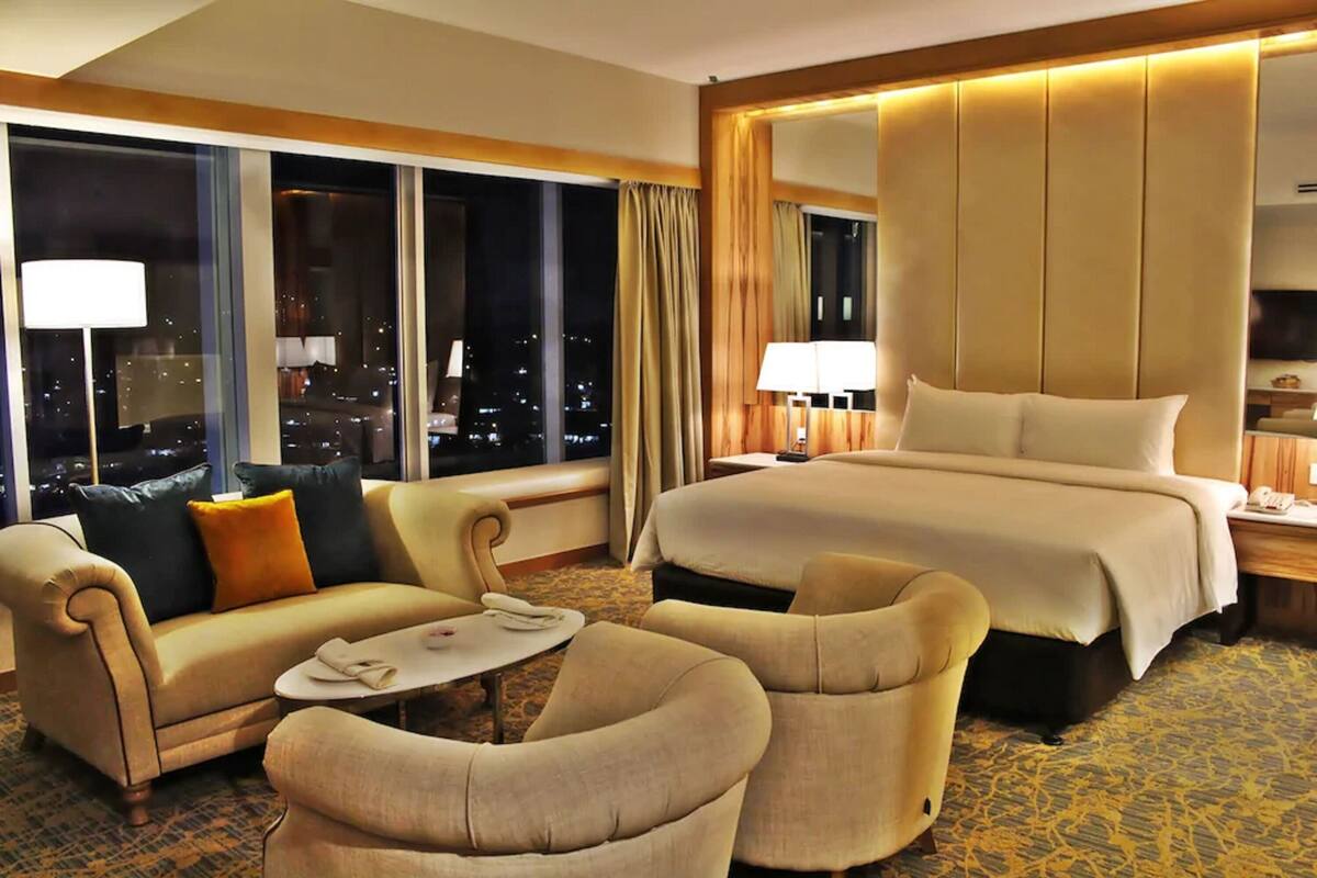 A hotel room with a large bed, a sofa, two armchairs, a small table, and a large window offering a city view.