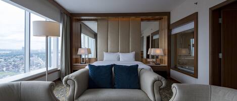 A hotel room with a large bed, two armchairs, a round table, and a city view.