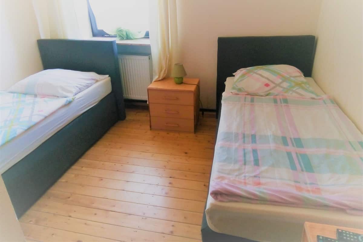 A small, simple room with two single beds, a wooden floor, a radiator, and a small table with a lamp.