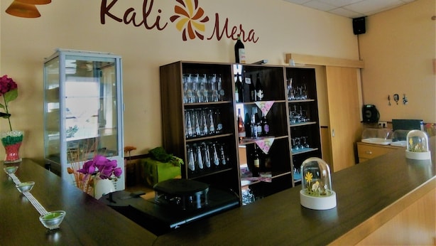 A reception area with a glass display case, a wooden counter, and shelves with wine bottles and glasses. - Hotel Kalimera (Heddesheim)