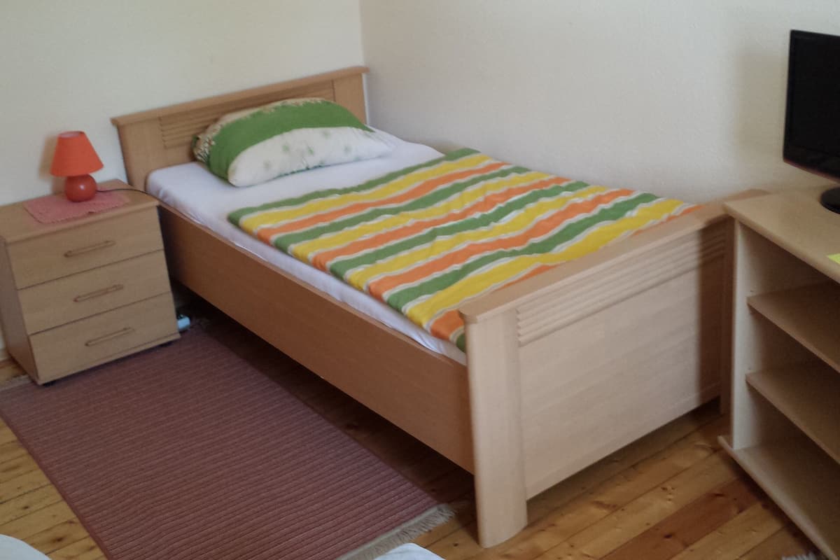 A single bed with a striped blanket, a nightstand with a lamp, a wooden dresser, and a television on a stand.