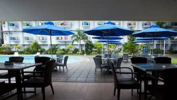 An outdoor dining area with tables and chairs, blue umbrellas, and a pool in the background.