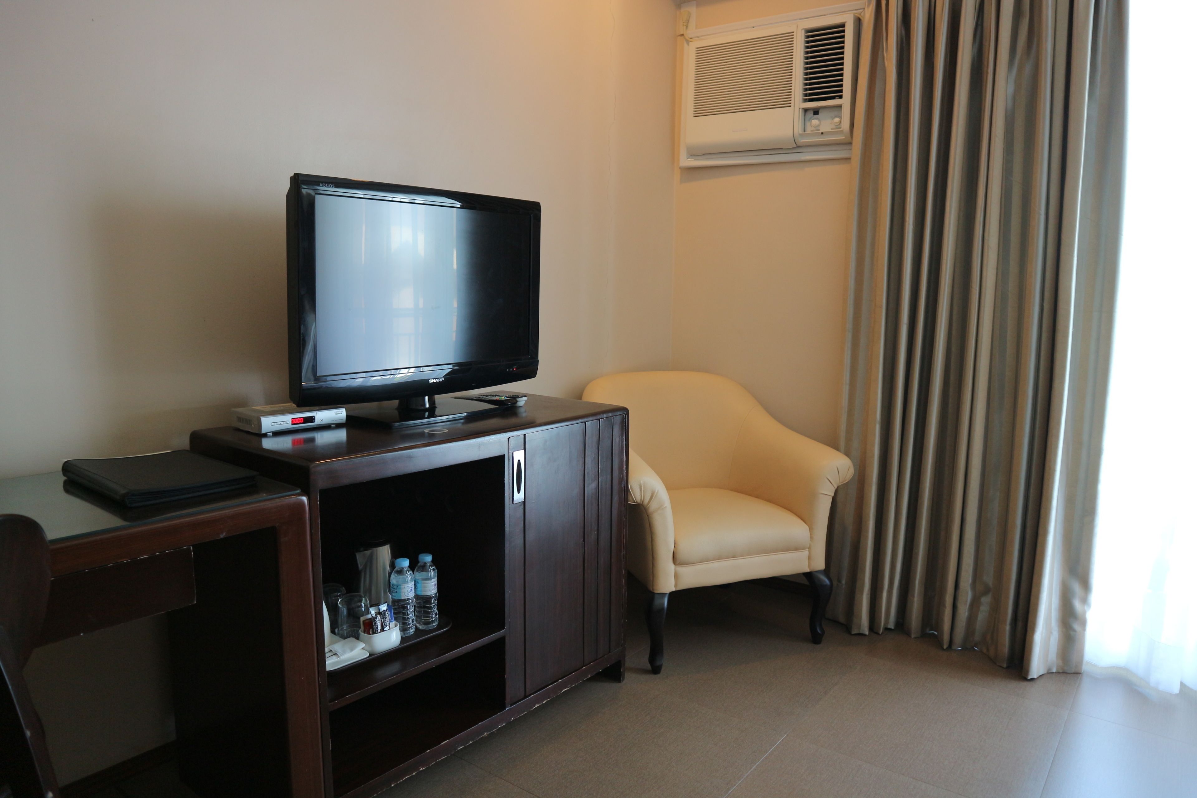 A room with a television on a wooden cabinet, a beige armchair, and a window with curtains.