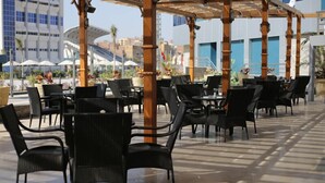 An outdoor patio with black wicker chairs and tables, a wooden pergola, and a view of buildings in the background.