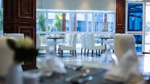 A restaurant with white chairs and tables, large windows, and a view of greenery outside.