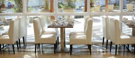 A restaurant with white chairs and tables, large windows, and potted plants.