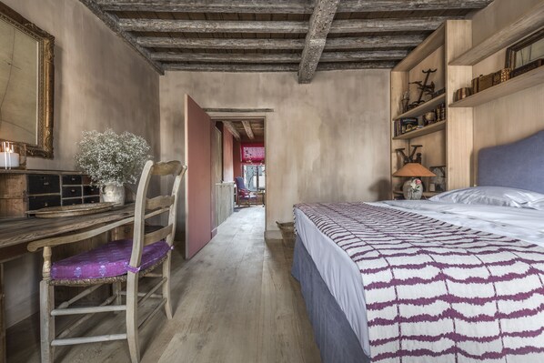 Ein Schlafzimmer mit einem Holztisch, einem Stuhl, einem Bett mit gemusterten Betttextilien und einem Regal mit verschiedenen Gegenständen.