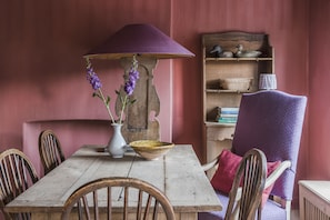 Una mesa de madera con un jarrón de flores moradas, un tazón y una lámpara morada. Hay sillas alrededor de la mesa y una estantería con objetos decorativos.