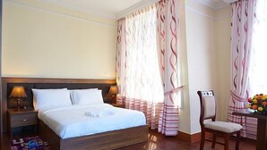 A neatly made bed with white linens, a wooden headboard, and a bedside table with a lamp. There is a chair with a cushion and a table with a vase of flowers. - Gentle Hotel (Addis Ababa)