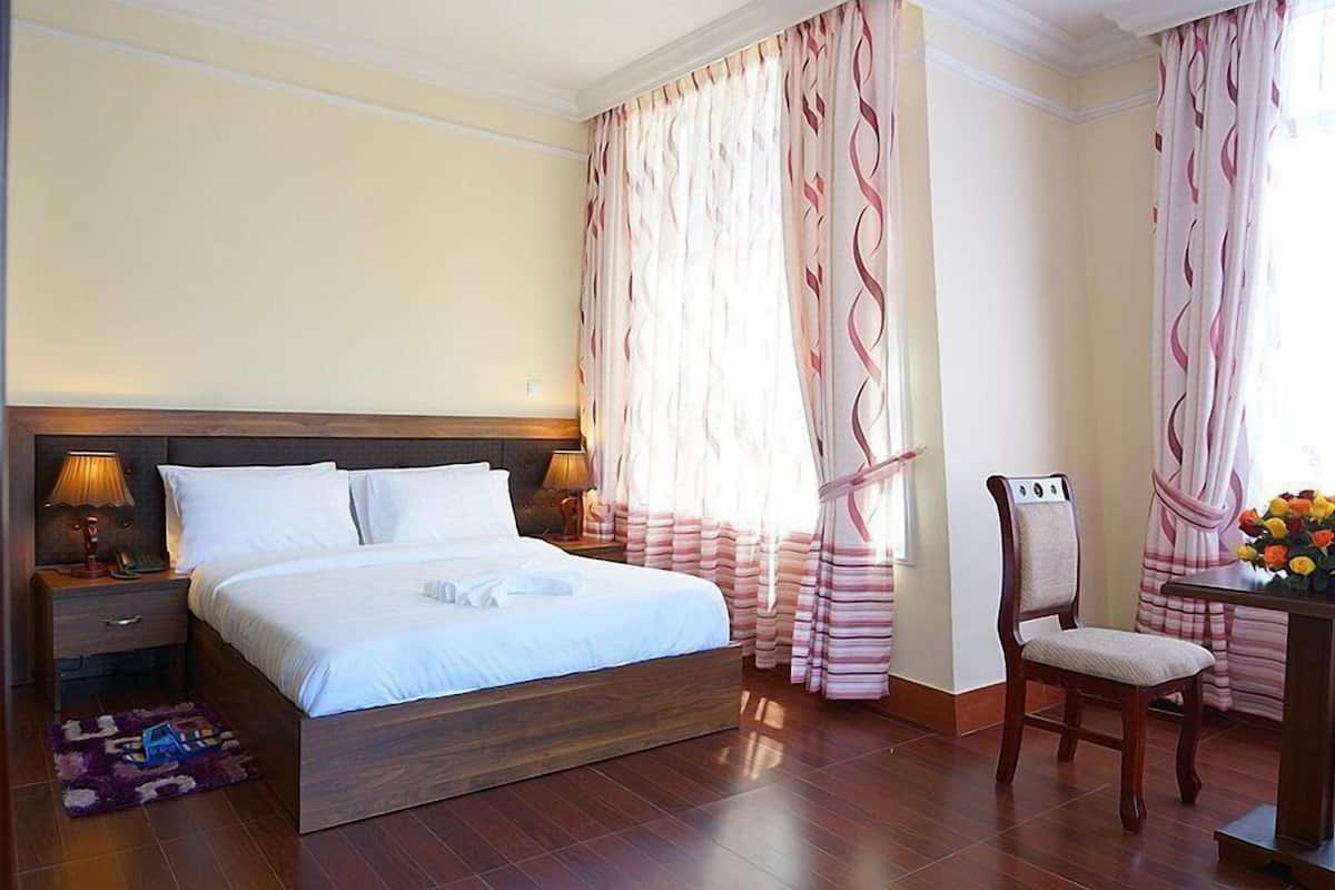 A neatly made bed with white linens, a wooden headboard, and a bedside table with a lamp. There is a chair with a cushion and a table with a vase of flowers.