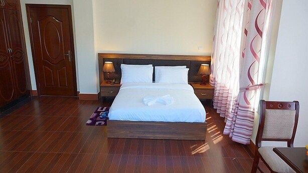 A neatly made bed with white linens, two bedside tables with lamps, a wooden headboard, and a patterned curtain. - Gentle Hotel (Addis Ababa)