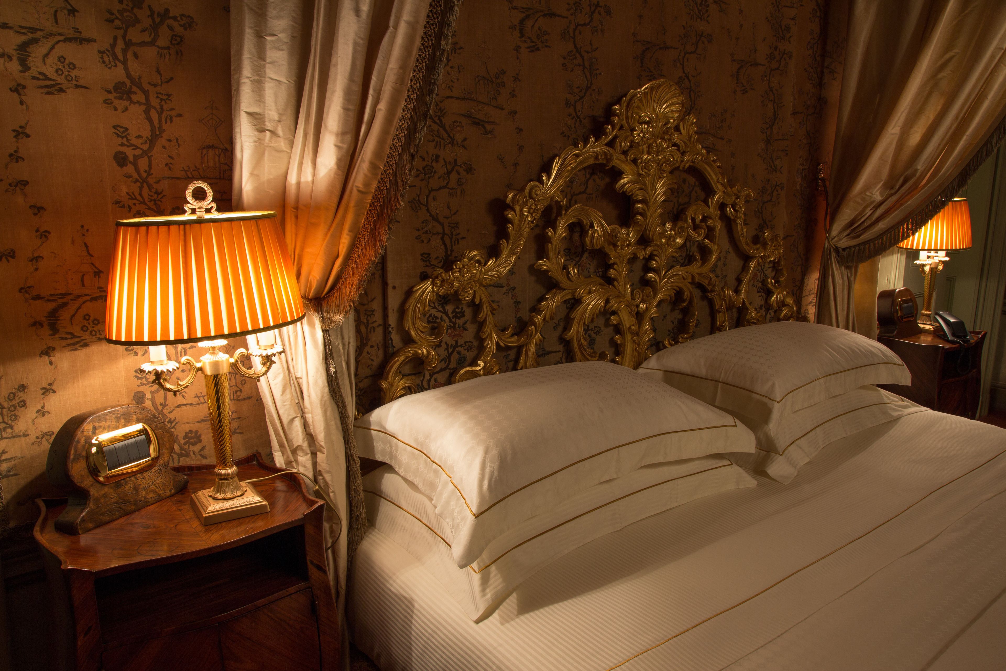 Ein luxuriöses Schlafzimmer mit einem aufwendig gestalteten Kopfteil, einer klassischen Nachttischlampe und einem ordentlich bezogenen Bett.