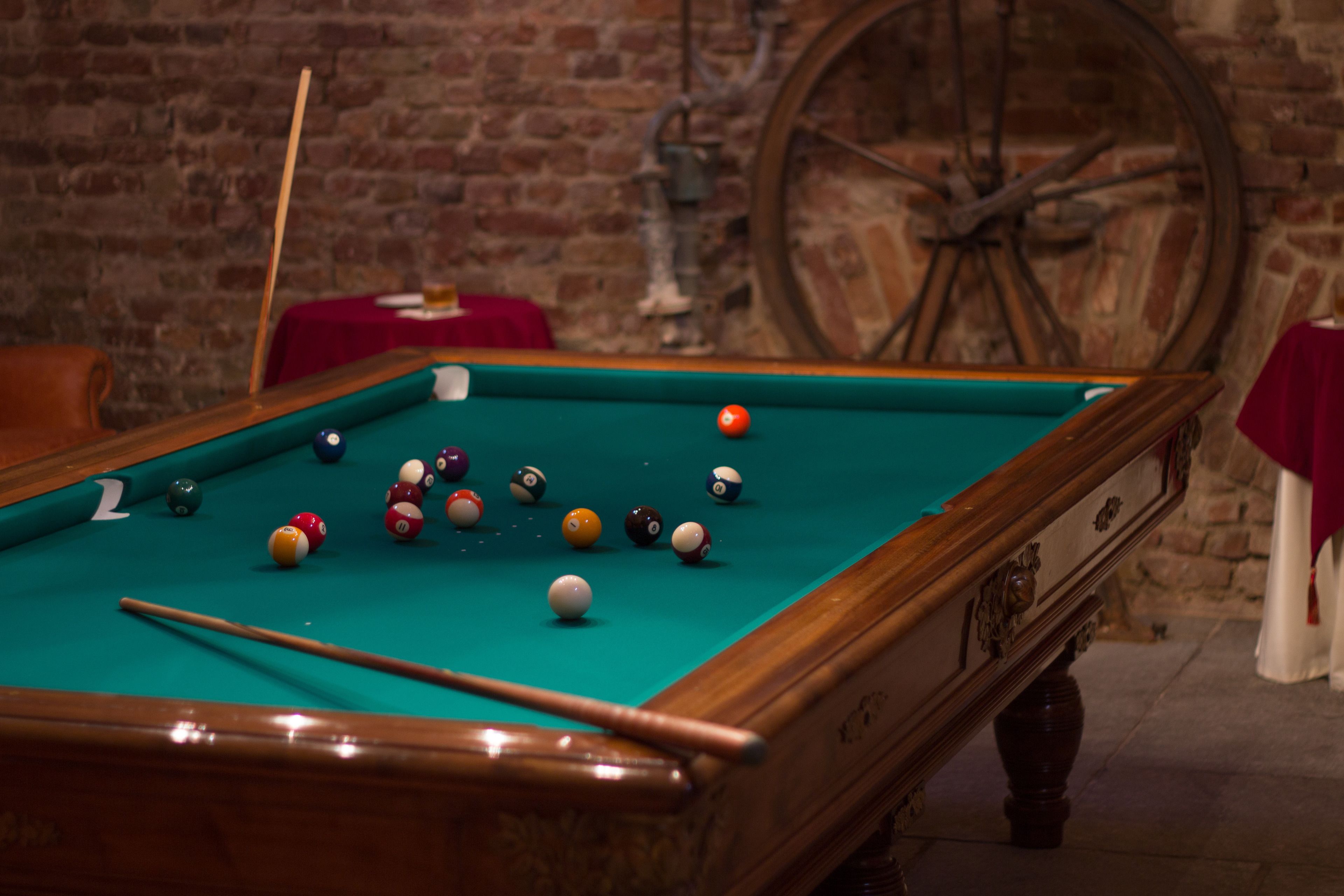 A pool table with balls, a wooden pool cue, and a brick wall with a large wooden wheel.