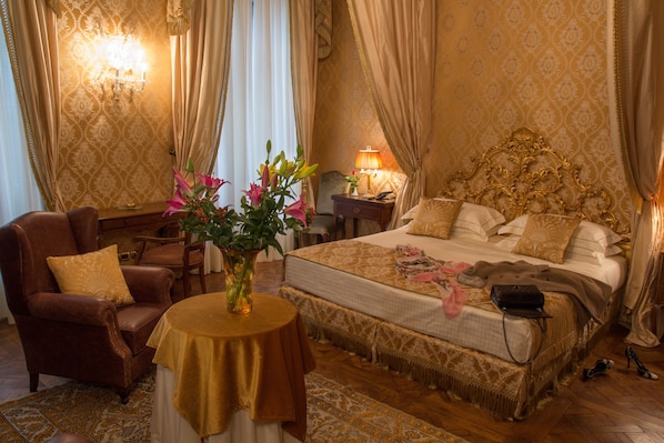 A luxurious bedroom with a large bed, a sitting area with a chair and a table, and a nightstand with a lamp. - Castello di Guarene (Guarene)
