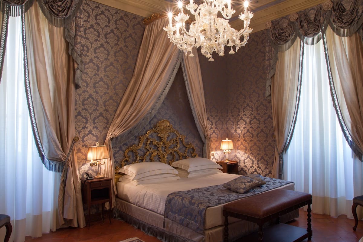 Ein Schlafzimmer mit einem großen Bett, einem verzierten Kopfteil und einer Chandelieber. Es gibt zwei Nachttische mit Lampen.