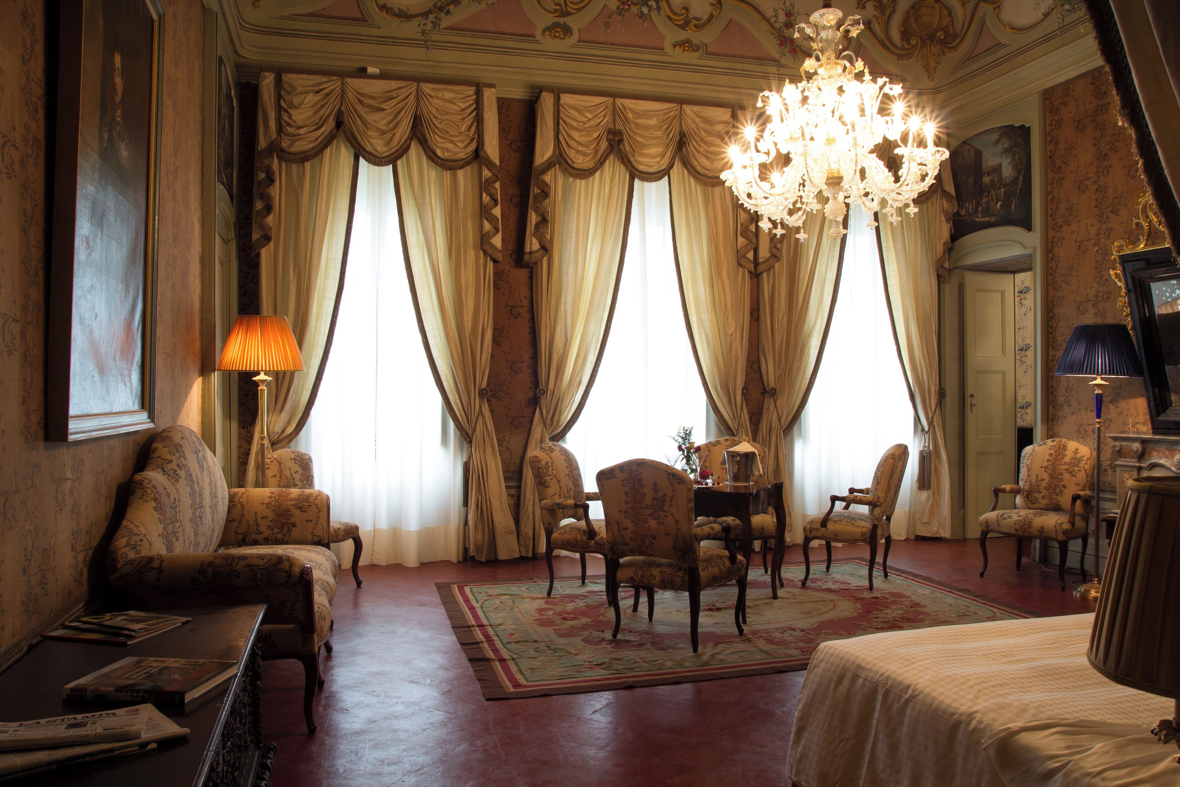 A room with a chandelier, patterned sofas, a dining table with chairs, and a large window with curtains.