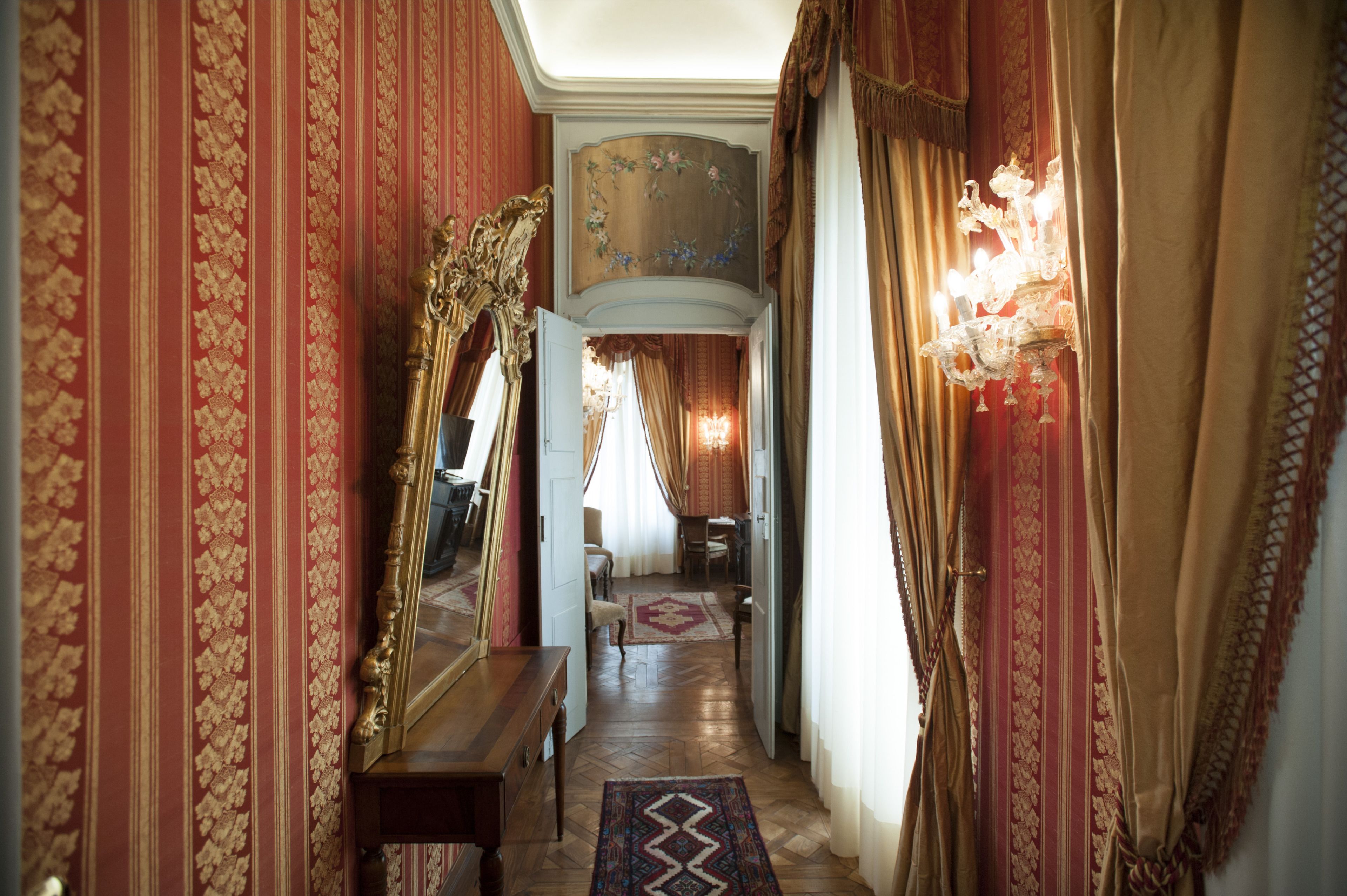A hallway with ornate red and gold wallpaper, a large mirror, and a chandelier.