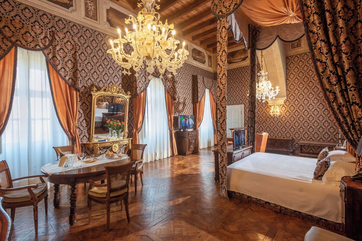 Ein Zimmer mit einem großen Bett, einer Kristallleuchter, einem Esstisch mit Stühlen und einem Spiegel.