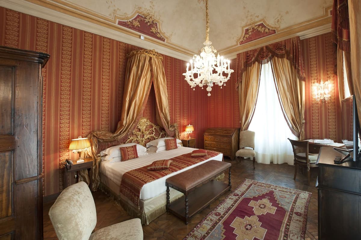 Ein geräumiges Schlafzimmer mit einem großen Bett, einer Kristalllüster, einem Schreibtisch mit Computer und einem klassischen Holzschrank.