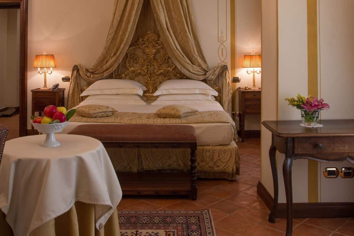 Ein Schlafzimmer mit Himmelbett, einer Kandelaberleuchter, einem Nachttisch mit Lampe, einem kleinen Tisch mit Obst und einem gemusterten Teppich.