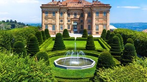 Um edifício grande e histórico com fachada simétrica, entrada central e um jardim formal com uma fonte.