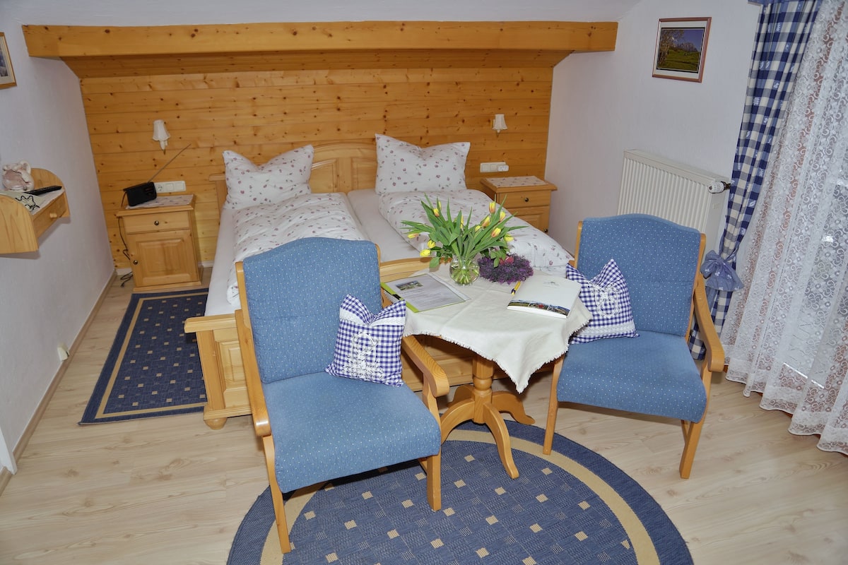 A bedroom with a wooden headboard, a double bed with floral bedding, two blue armchairs, a small table with a vase of flowers, and a nightstand with a lamp.