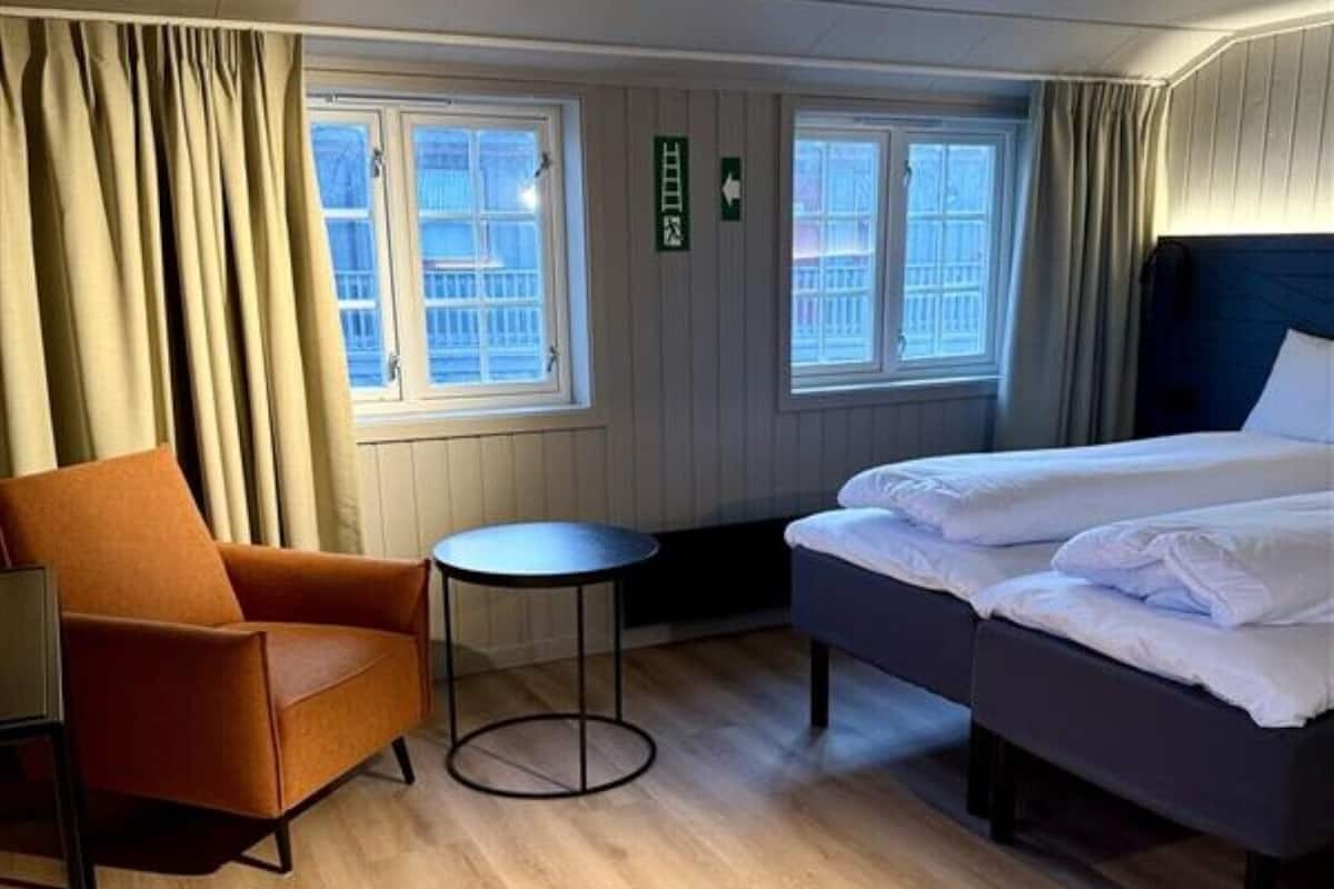 Quarto de hotel com cama, cadeira, mesa pequena e janelas com cortinas.