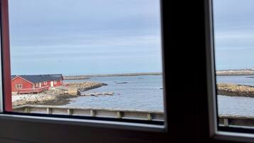 A view through a window of a wooden deck leading to a rocky shoreline with a red building.