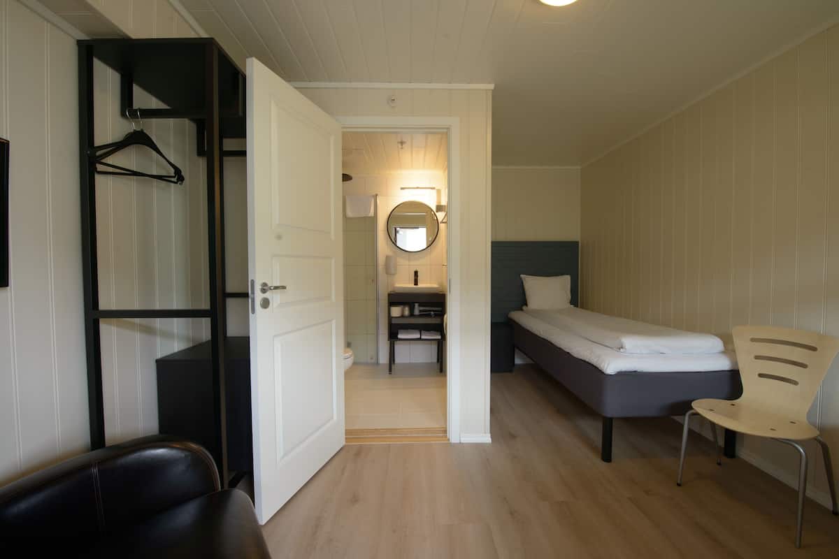 Quarto pequeno com cama, cadeira, cabideiro e banheiro visível por uma porta aberta.