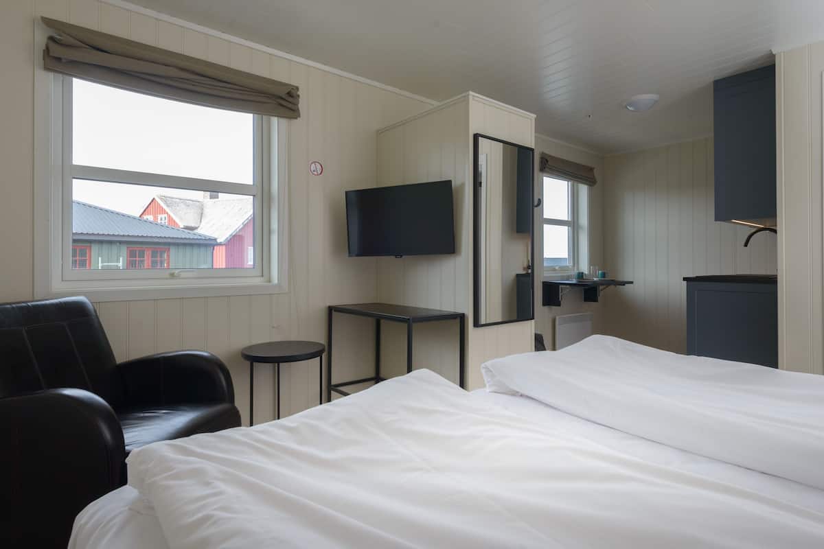 Quarto de hotel com uma cama, uma cadeira, uma televisão, uma mesinha e uma janela com vista para edifícios.
