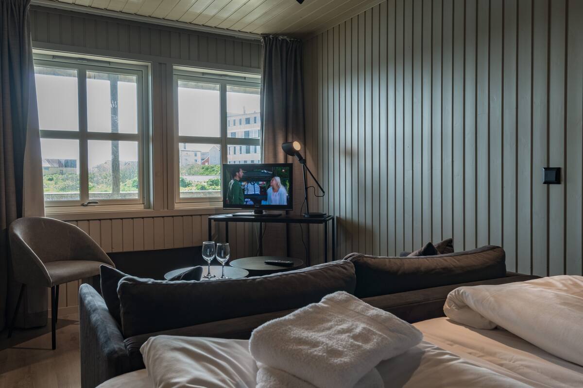 Quarto de hotel com cama, sofá, televisão e mesa com dois copos.