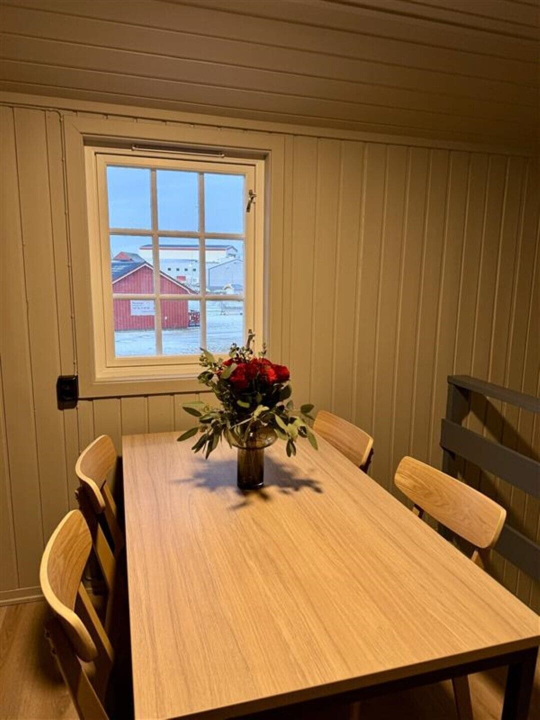 Un comedor con una mesa y sillas de madera, una ventana con vista a un edificio rojo, y un jarrón con flores rojas.