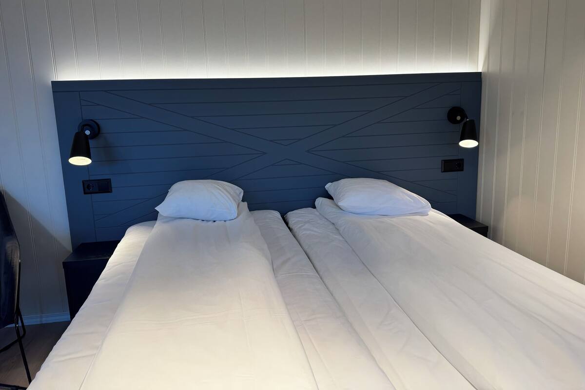 Cama de casal com lençóis brancos, duas luminárias de cabeceira e cabeceira azul com padrão geométrico.