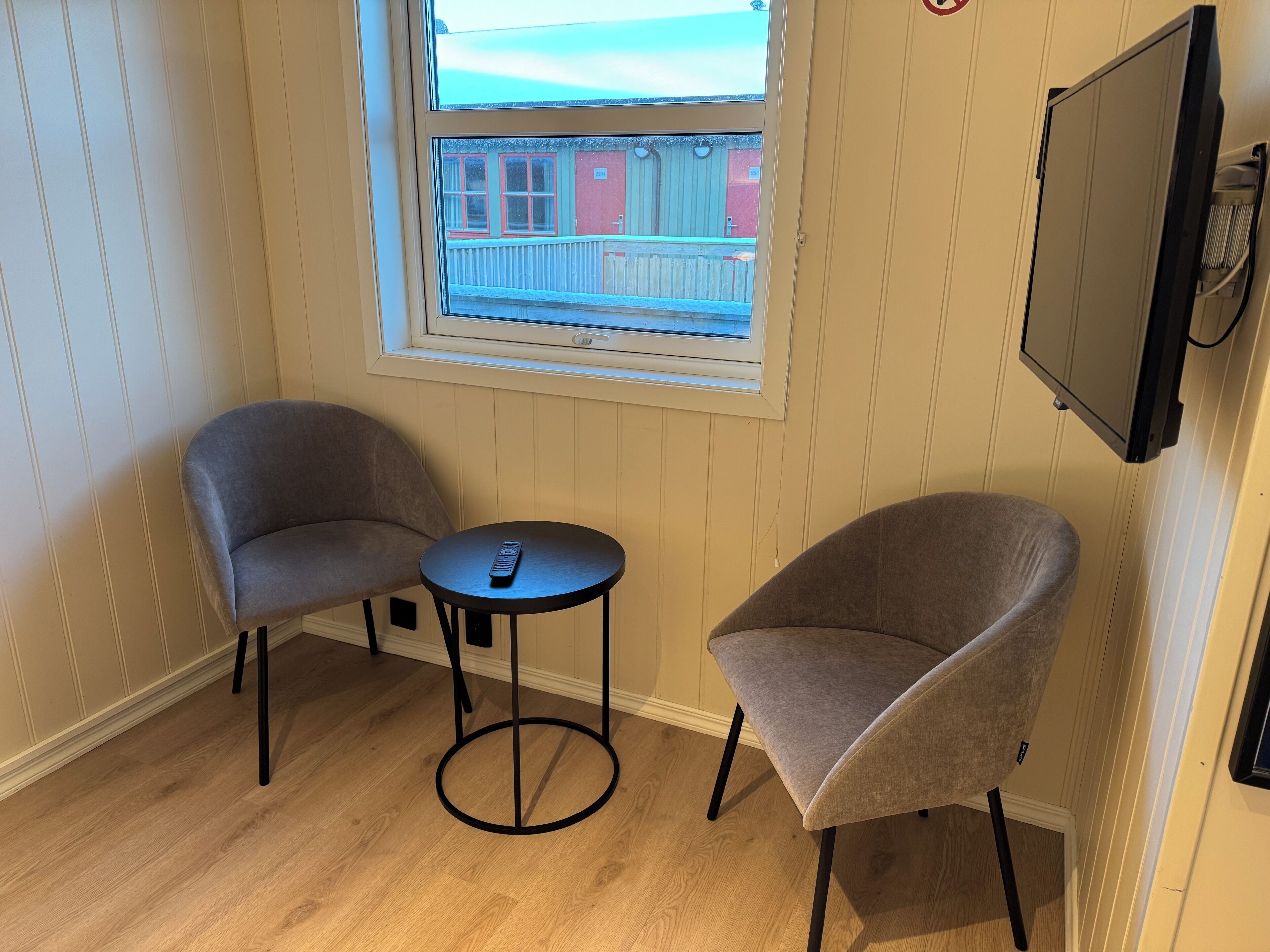 A room with two grey armchairs, a small round table with a remote control, and a TV mounted on the wall.