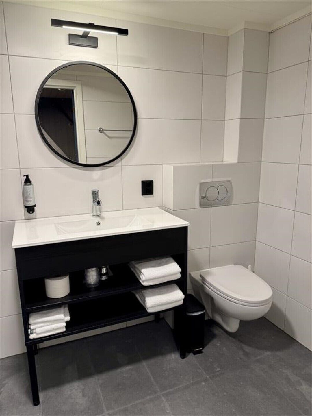 Een moderne badkamer met een wit wastafel, zwart badmeubel, ronde spiegel, toilet en handdoekhouder.