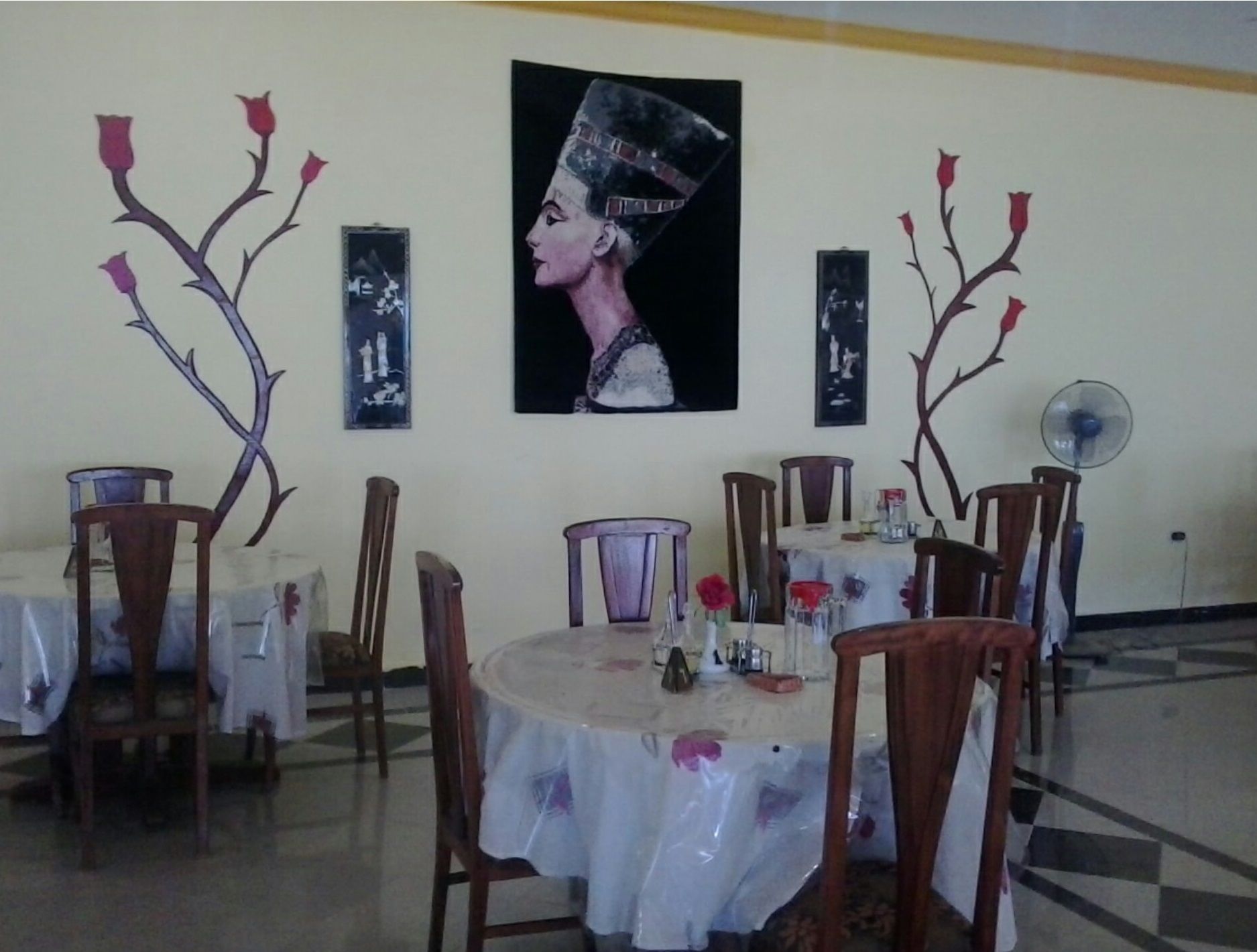 A dining area with round tables covered in white tablecloths, wooden chairs, and a central painting of a woman in a headdress.