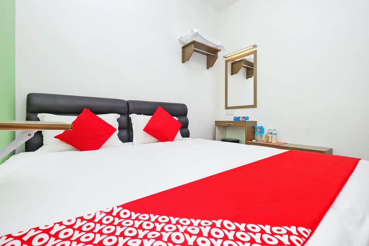 Una cama bien tendida con una colcha blanca y roja, un cabecero negro y dos almohadas rojas. Hay una mesita de noche pequeña con un espejo encima y algunos artículos de tocador.
