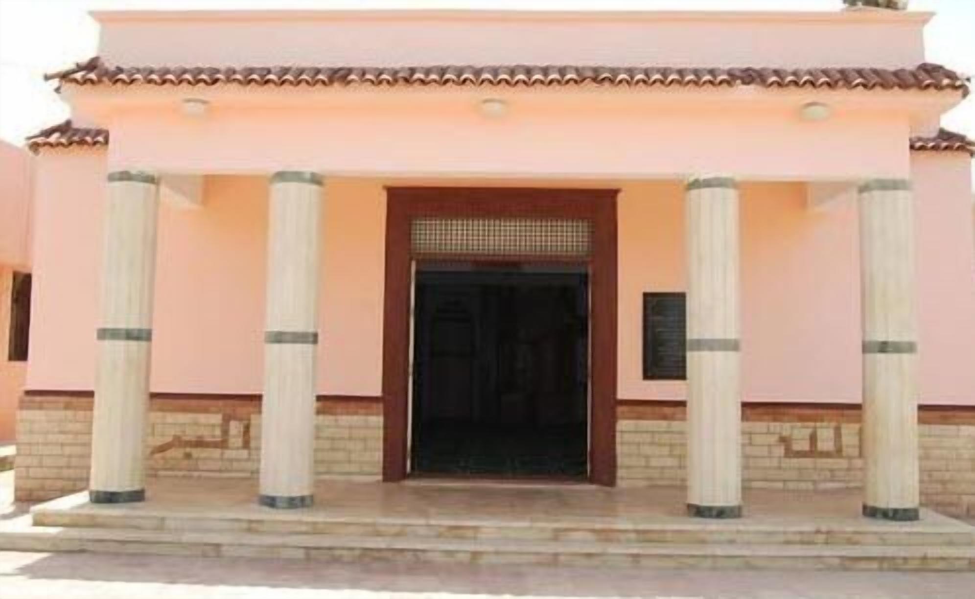 A building with a pink facade, a central entrance with a dark door, and four columns supporting the entrance.