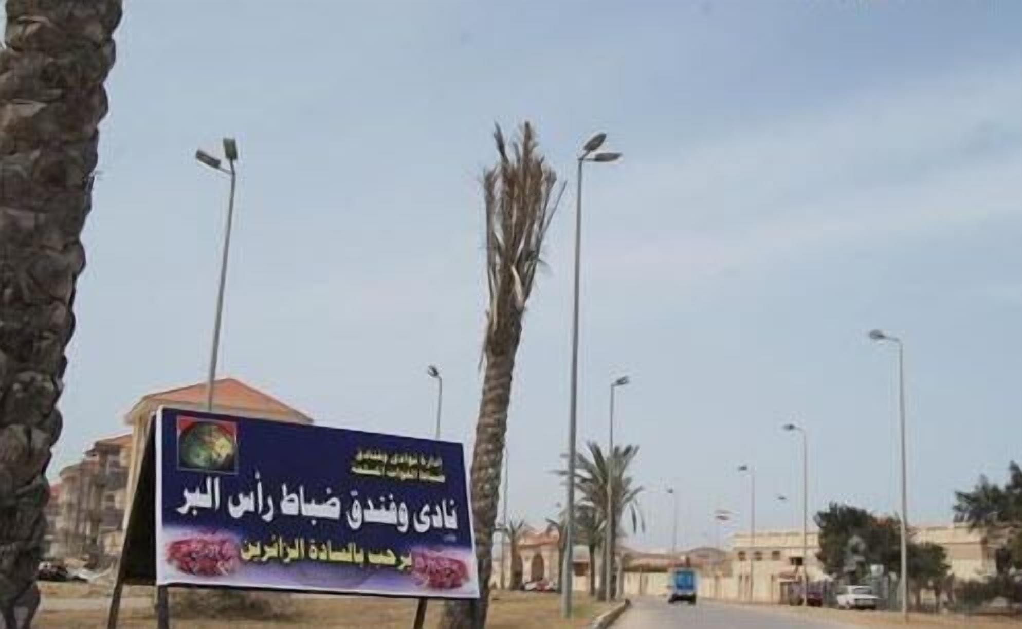 A signboard with Arabic text, palm trees, and streetlights along a road.