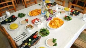 Et spisebord dækket med forskellige retter, herunder fisk, salater og ris, i et rum med træstole.