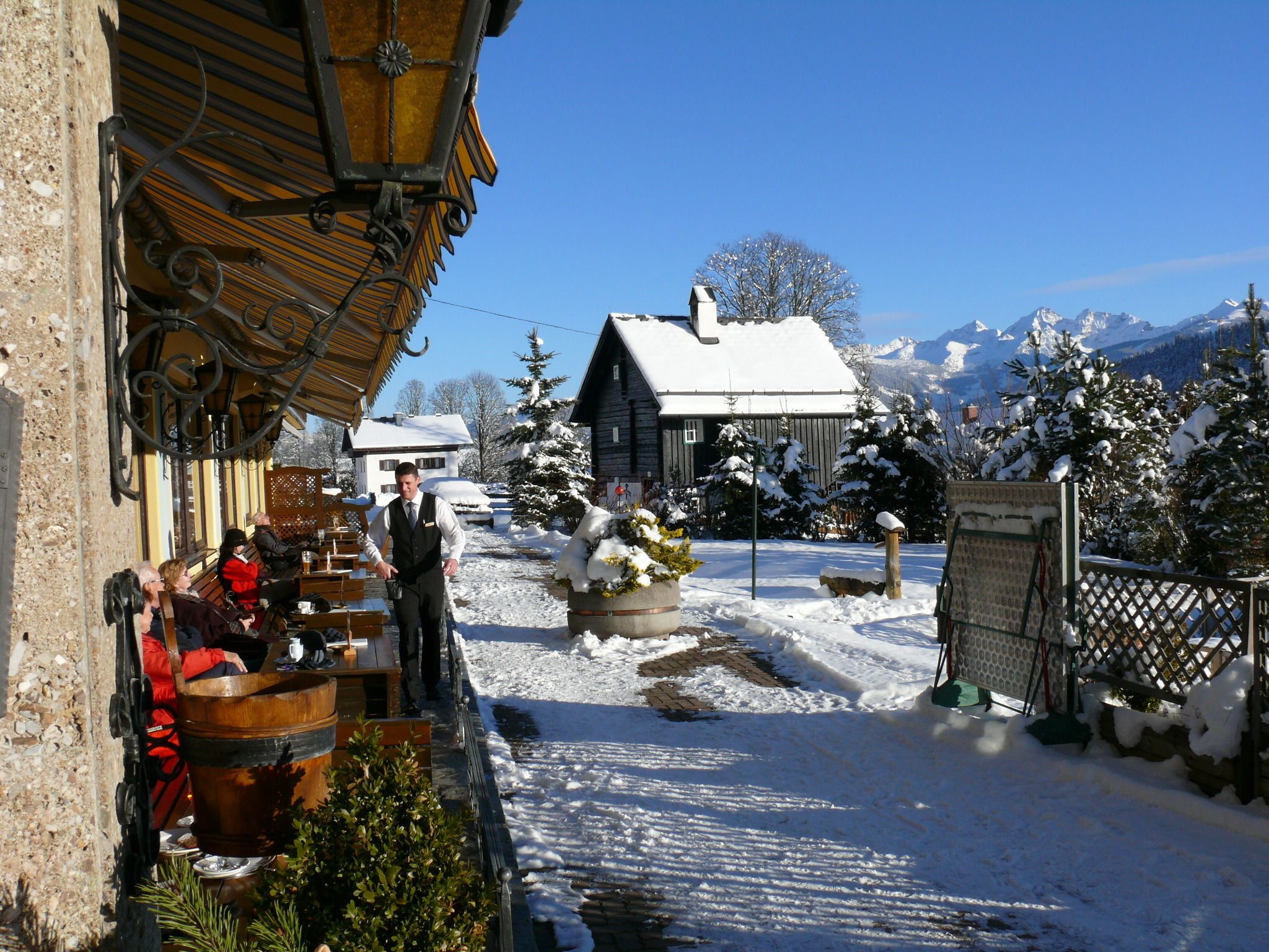 A snowy outdoor area with a wooden building, a snow-covered tree, and a mountain in the background.