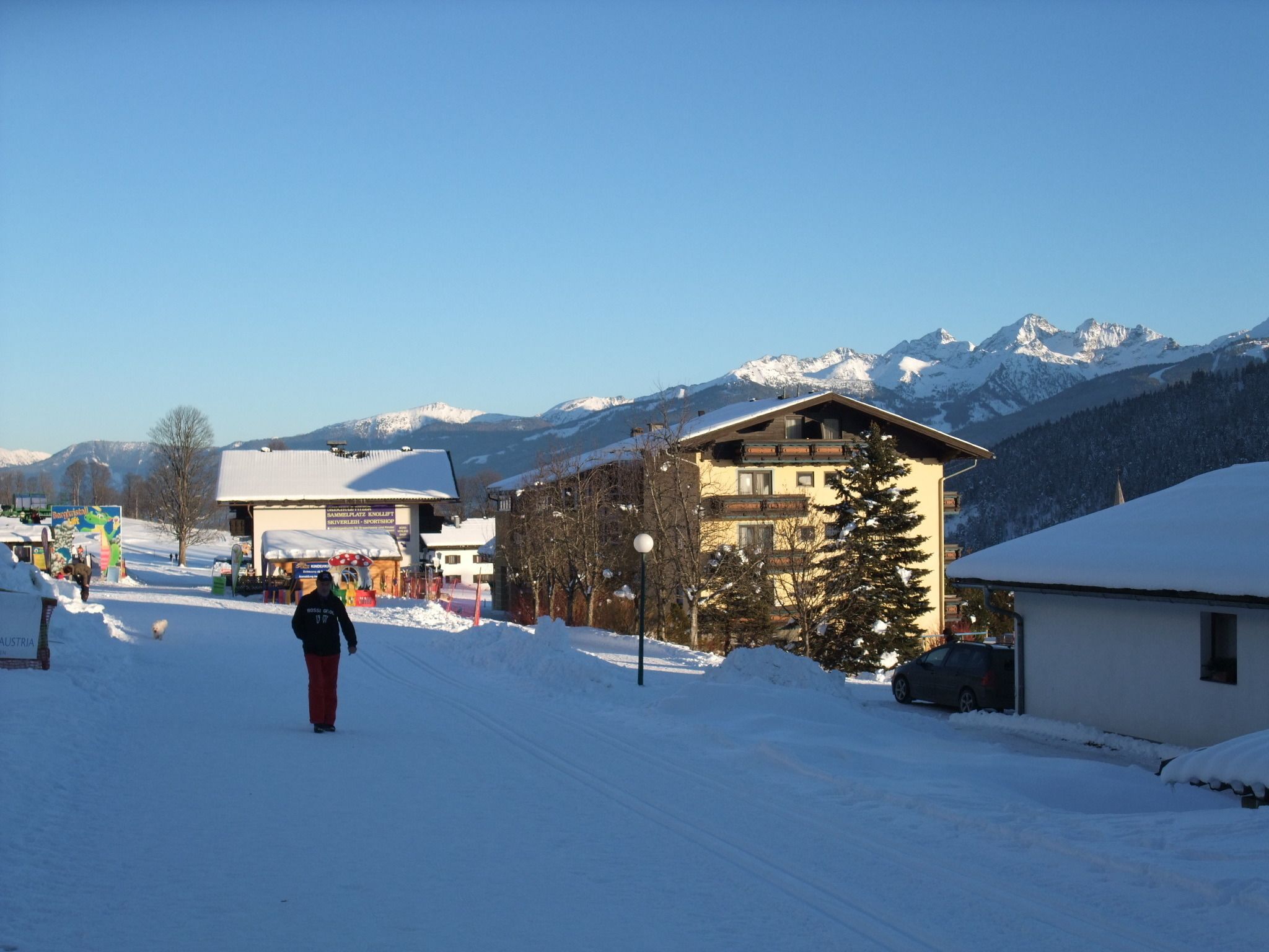 A snow-covered street with buildings, a person walking, and mountains in the background.