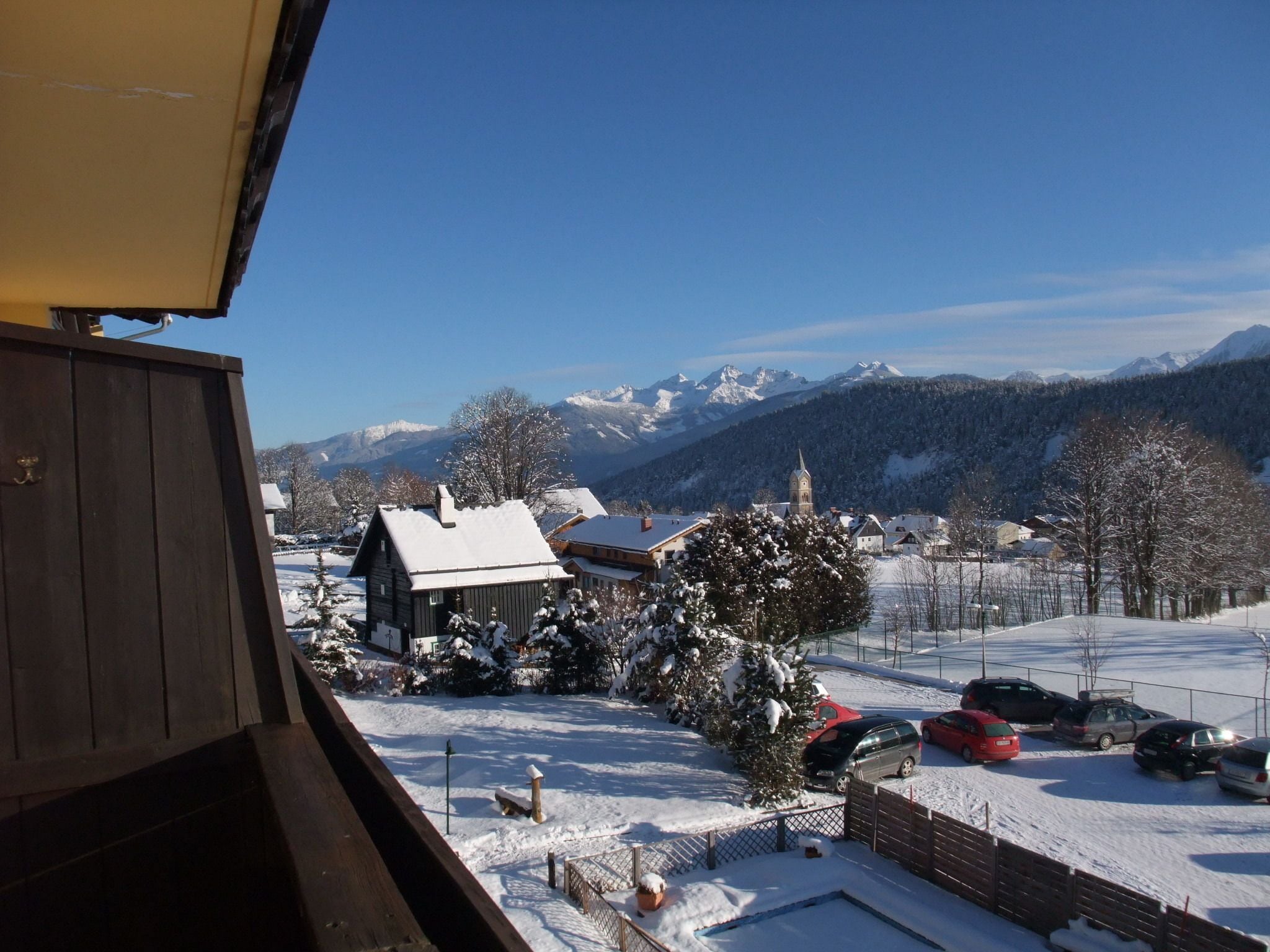 A snowy mountain village with a church, residential buildings, and parked cars.