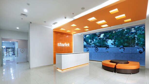 A hotel reception area with a modern design, featuring a reception desk, seating area, and a large window with a view of trees. - Starlet Hotel Serpong (South Tangerang)