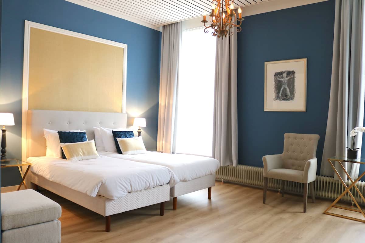 Una habitación de hotel con una cama grande, dos mesitas de noche, una silla, una mesita con un jarrón y un cuadro abstracto de gran tamaño en la pared.