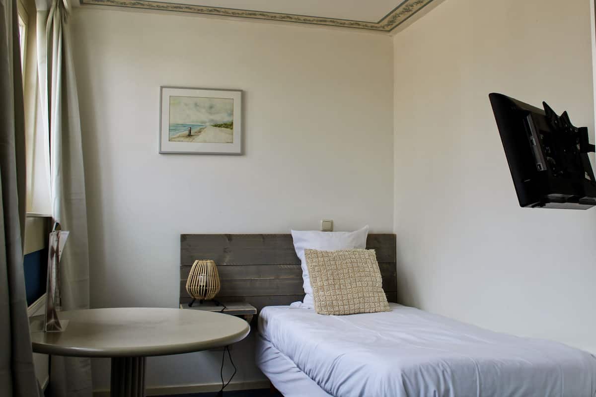 Un dormitorio con cama, mesita de noche, lámpara, un cuadro en la pared y televisor montado en la pared.