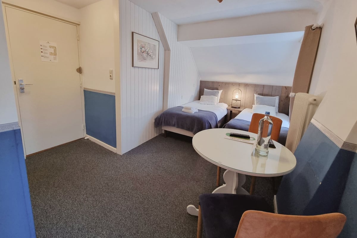 Habitación de hotel con dos camas, una mesita redonda pequeña, una silla y una puerta.