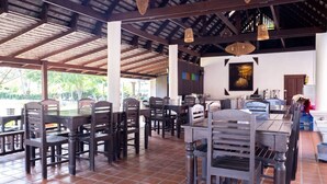 Un amplio espacio exterior para comer con mesas y sillas de madera, suelo de baldosas y techo cubierto con luces colgantes.