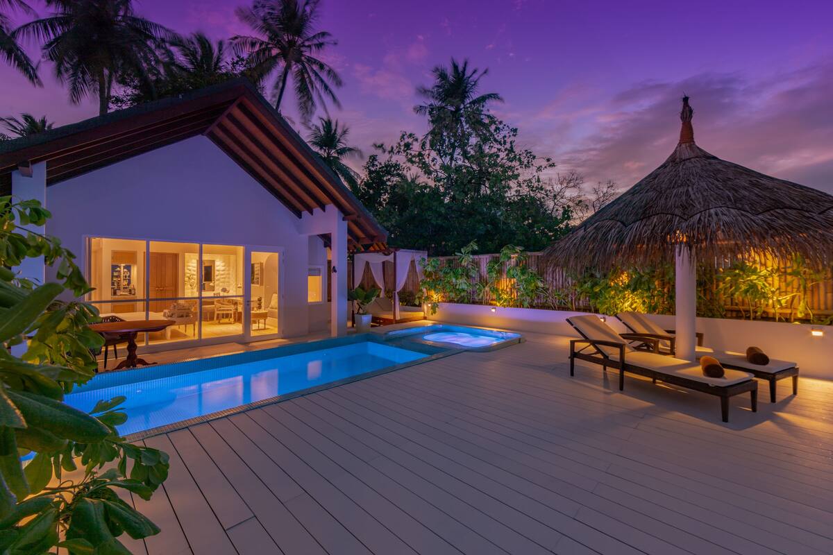 A poolside area with a house, lounge chairs, and a thatched-roof structure.